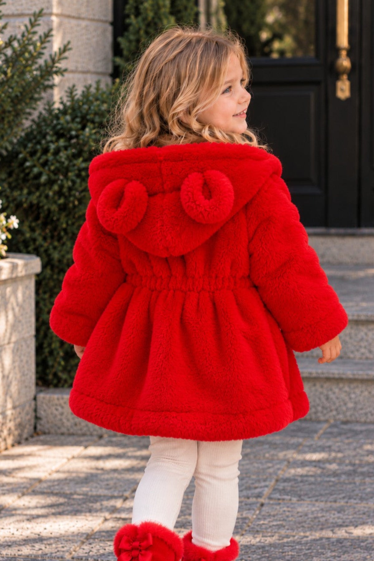 A young girl wearing a bright red plush hooded coat with two pom-poms on the hood, white leggings, and red fuzzy boots, standing on stone steps outdoors.