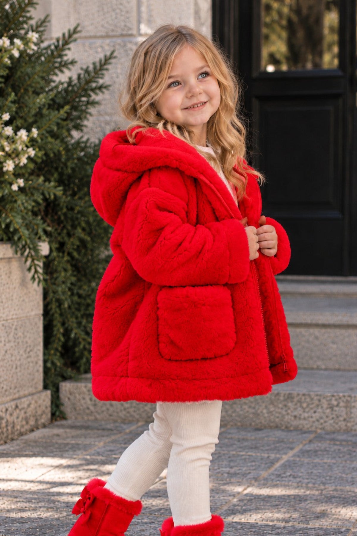 A young girl wearing a bright red plush hooded coat, white leggings, and red bow-front ankle boots, standing on stone steps outdoors.