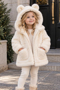 A young girl wearing a cream-colored faux fur hooded jacket with bear ears on the hood, standing on stone steps in front of a building.