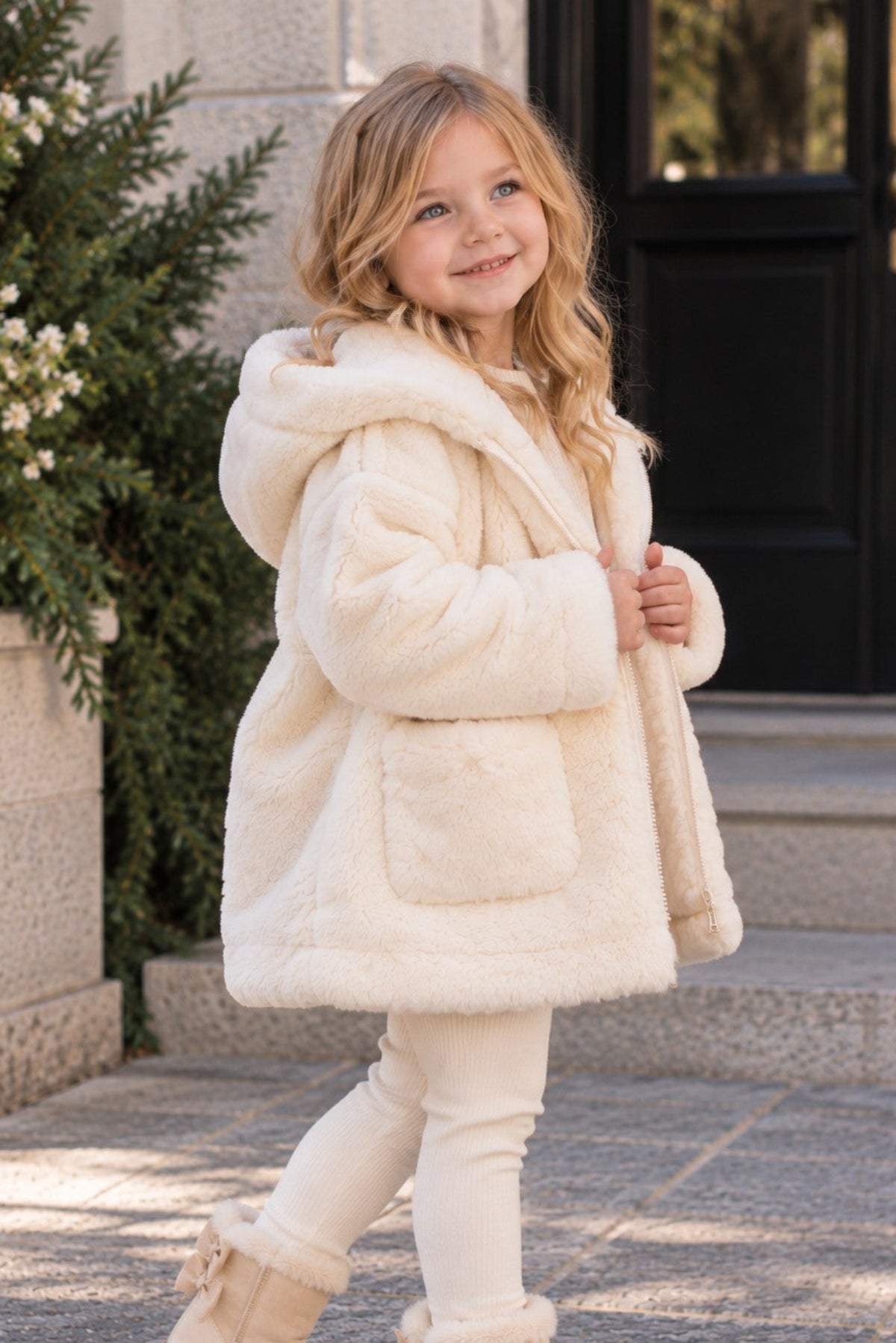 A young girl wearing a fluffy cream faux-fur hooded coat, cream leggings, and beige ankle boots, standing on a stone walkway in front of a building.