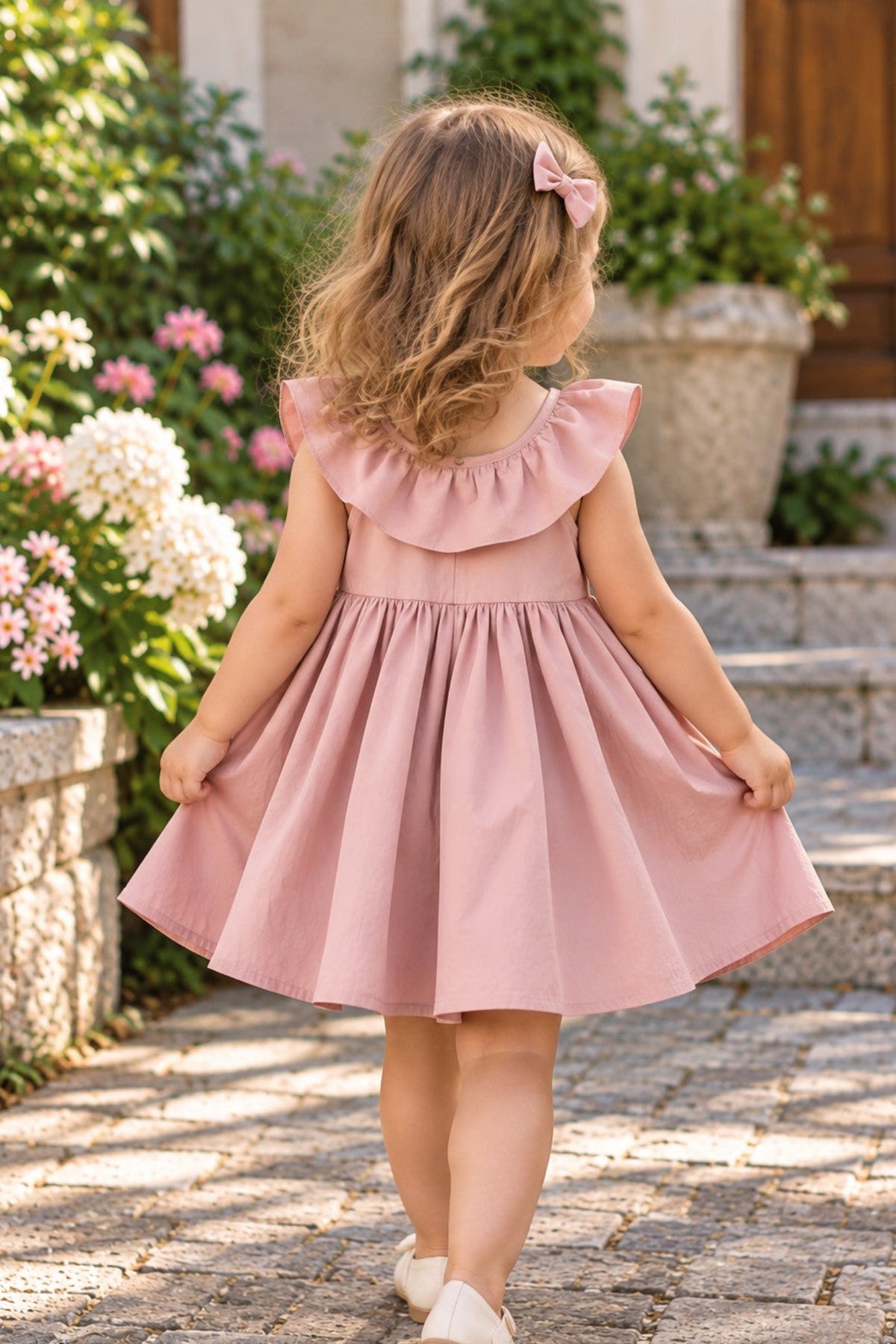 A young girl with blonde hair wearing a dusty pink sleeveless dress with a ruffled collar and a matching hair bow, walking away on a stone path in a garden setting.