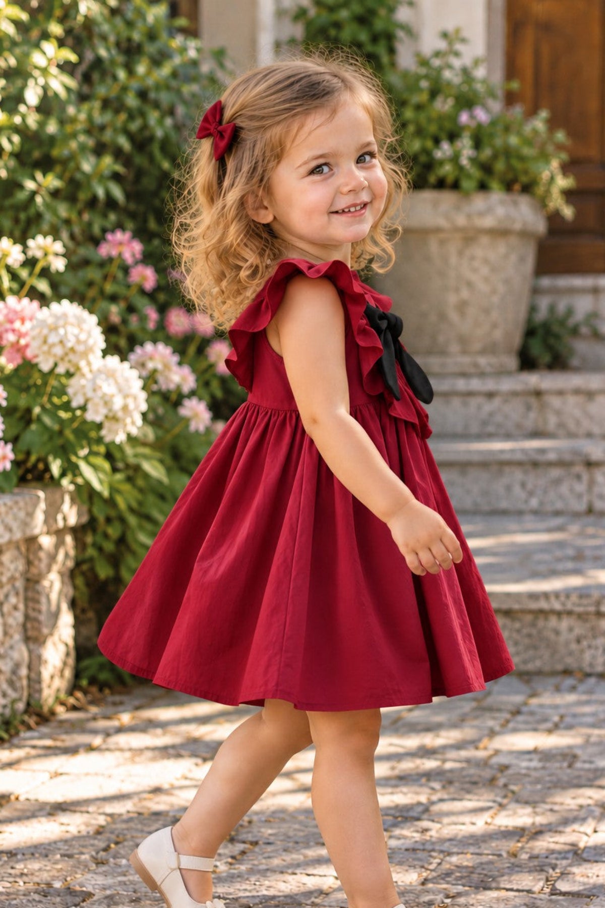 A young girl with curly blonde hair wearing a sleeveless deep red dress with ruffled shoulders and a black bow, walking outdoors on a stone path with flowers and steps in the background.