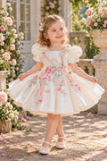 A young girl stands outdoors in a garden courtyard wearing a white or cream formal party dress with pink floral embroidery, puffed short sleeves, and a full skirt. She is holding out both sides of the skirt to show its volume, with stone planters, flowers, and a building facade in the background.