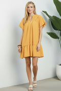 Woman wearing a yellow dress standing indoors with a plant in the background