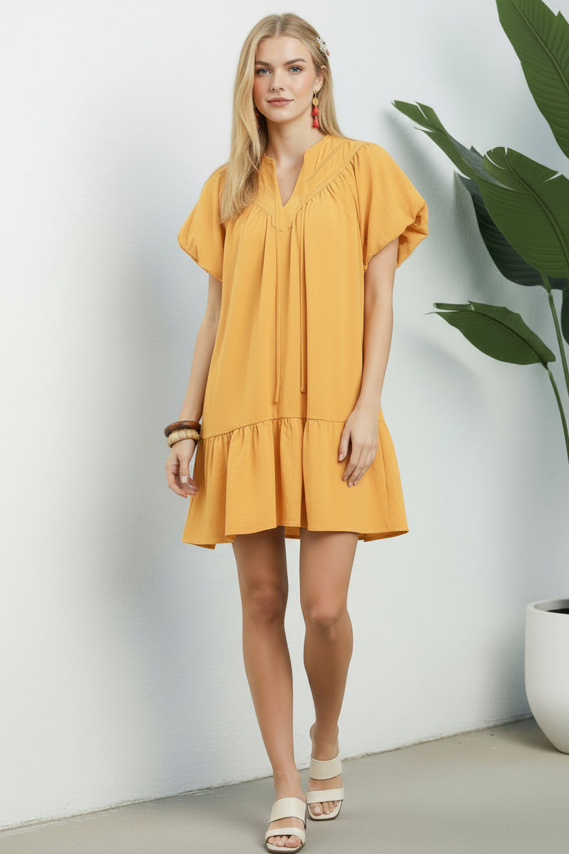 Woman wearing a yellow dress standing indoors with a plant in the background