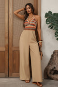 Woman wearing a patterned top and beige jumpsuit standing against a wooden wall with a plant.
