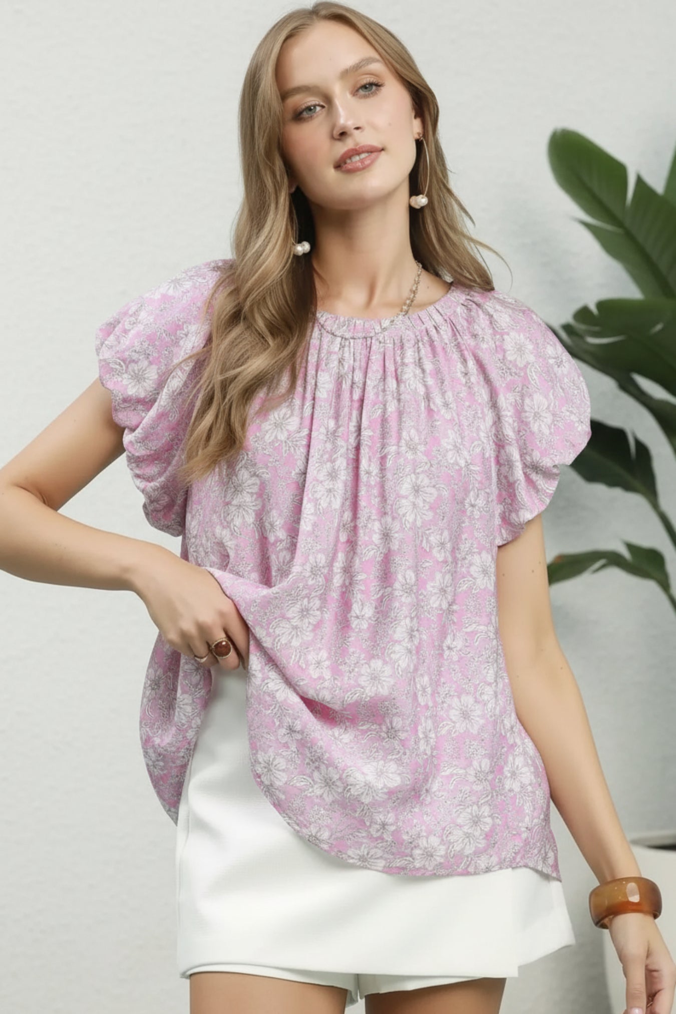 Woman wearing a pink floral blouse with white shorts on a white background with plant