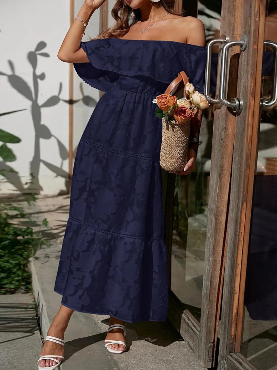 Woman in a navy off-shoulder dress holding a floral basket, standing in front of a wooden door.