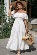 Woman in a white off-shoulder dress with a straw hat and bag, standing outdoors.