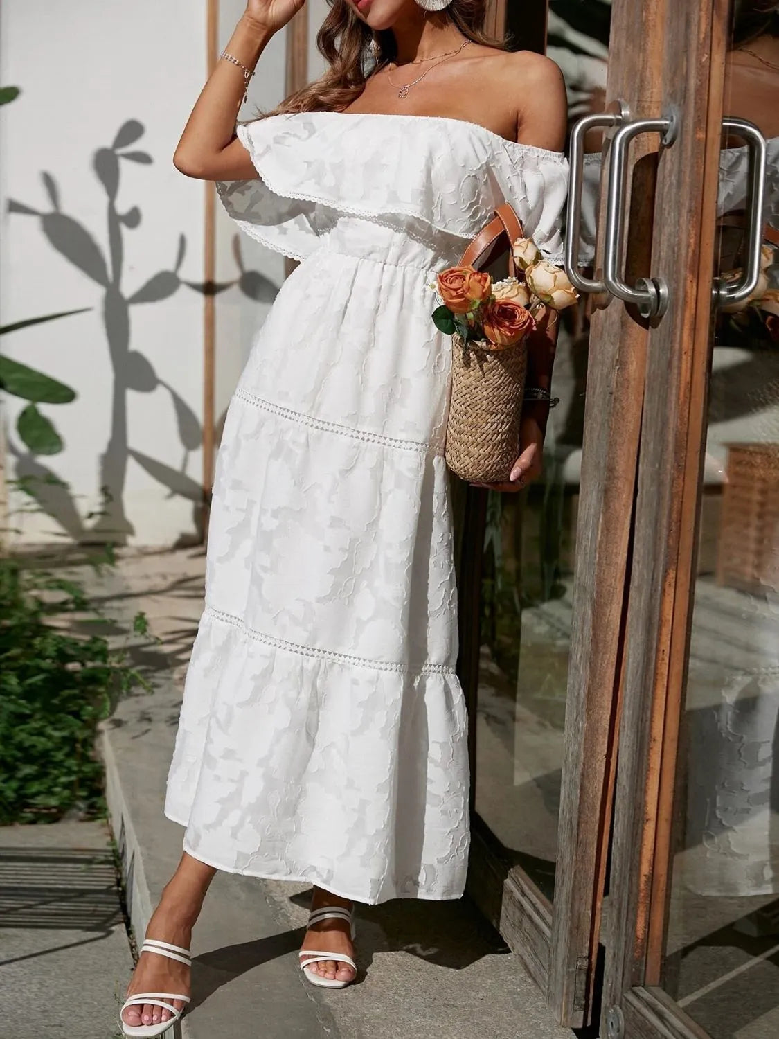 Woman in a white off-shoulder dress holding flowers, standing by a door.