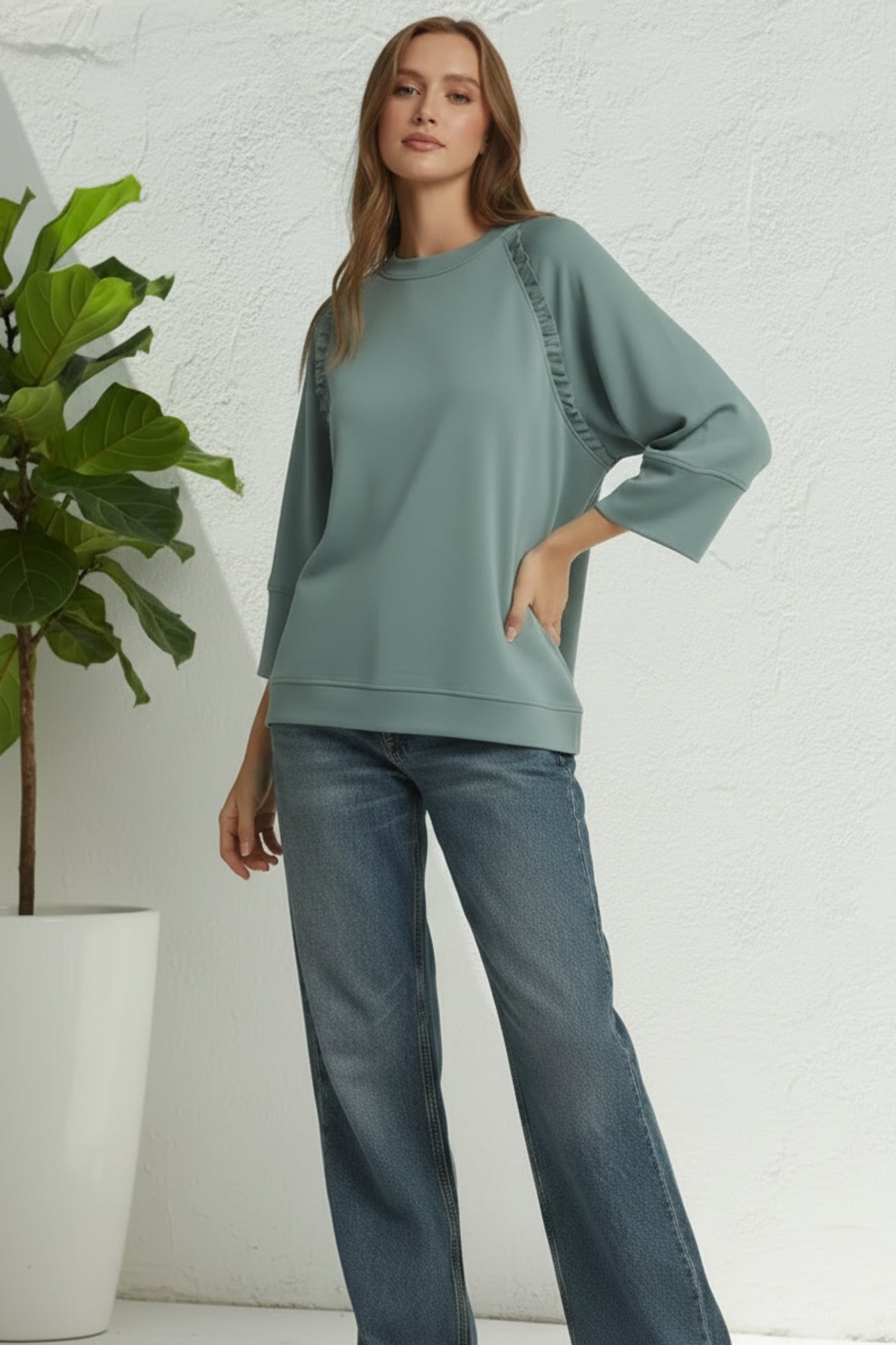 Woman wearing a green sweater with blue jean pants against a white background with plant on the side