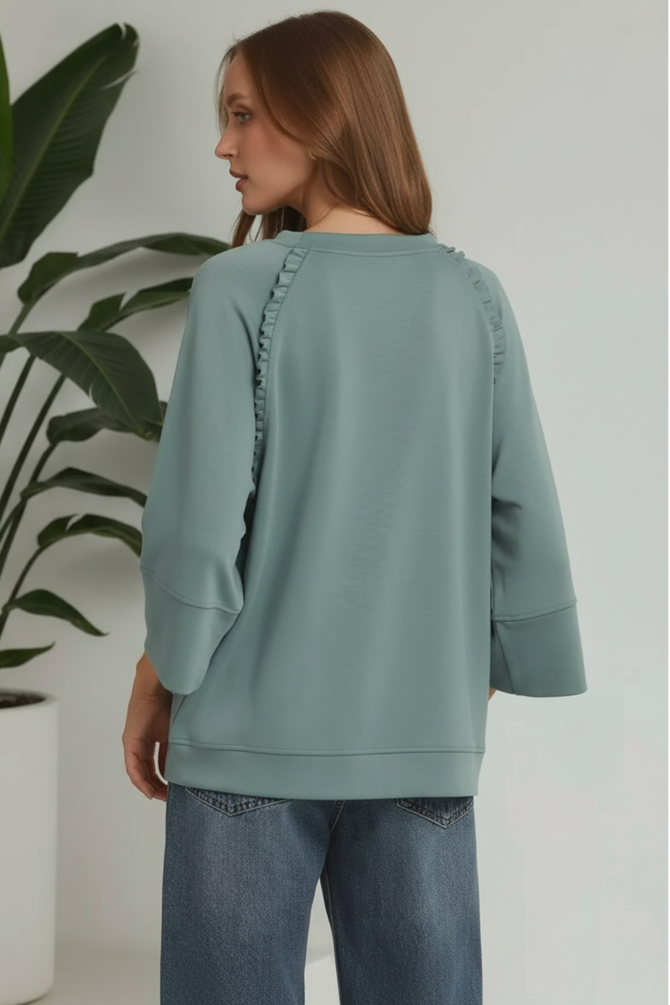Person wearing a light green sweater with ruched details on a white background with plant on the side
