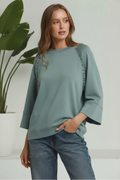 Woman wearing a green sweater with raglan sleeves against a white background with plant on the side