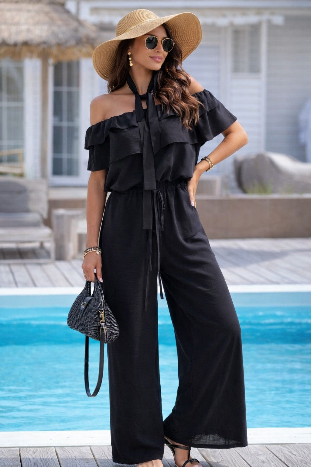 Woman in a black off-shoulder jumpsuit with a wide-brimmed hat by a pool.