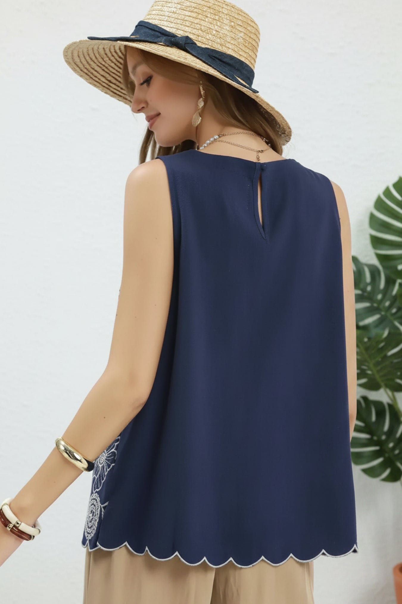 Woman wearing a navy sleeveless top with scalloped hem and a wide-brimmed hat on a light gray background.