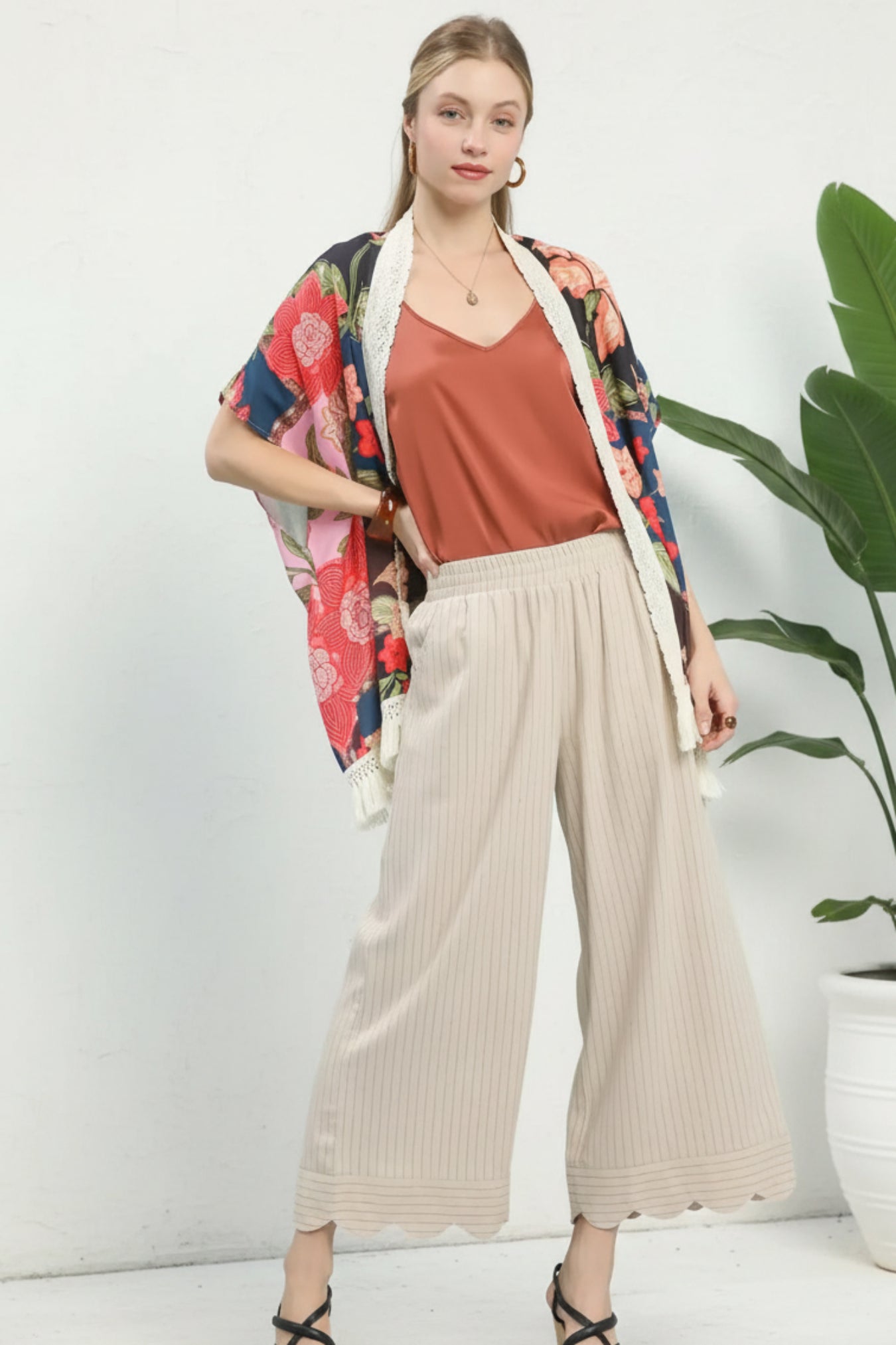 Woman wearing a colorful kimono-style jacket, rust top, beige wide-leg pants, and black sandals on a white background.