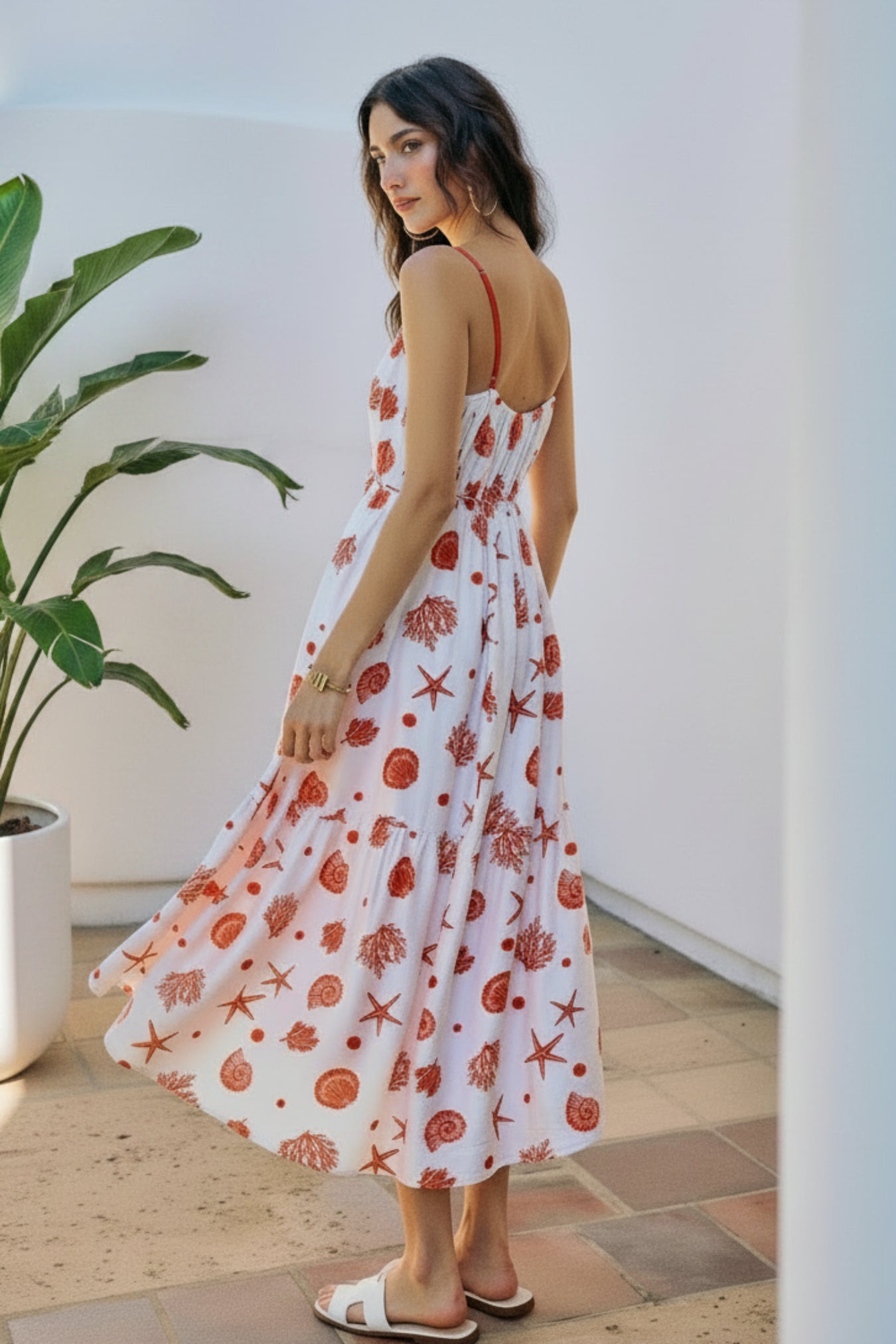 Woman in a white dress with red patterns standing in a sunlit outdoor setting with a plant.