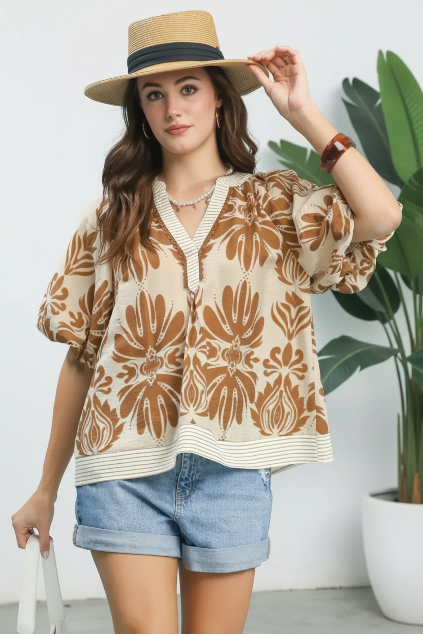 Woman wearing a patterned blouse, denim shorts, and a straw hat against a white background.