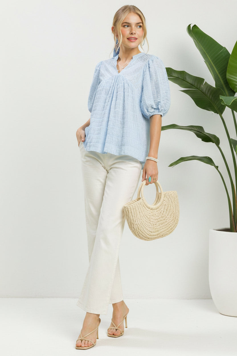 Woman wearing a light blue blouse and white pants, holding a straw bag on a green background