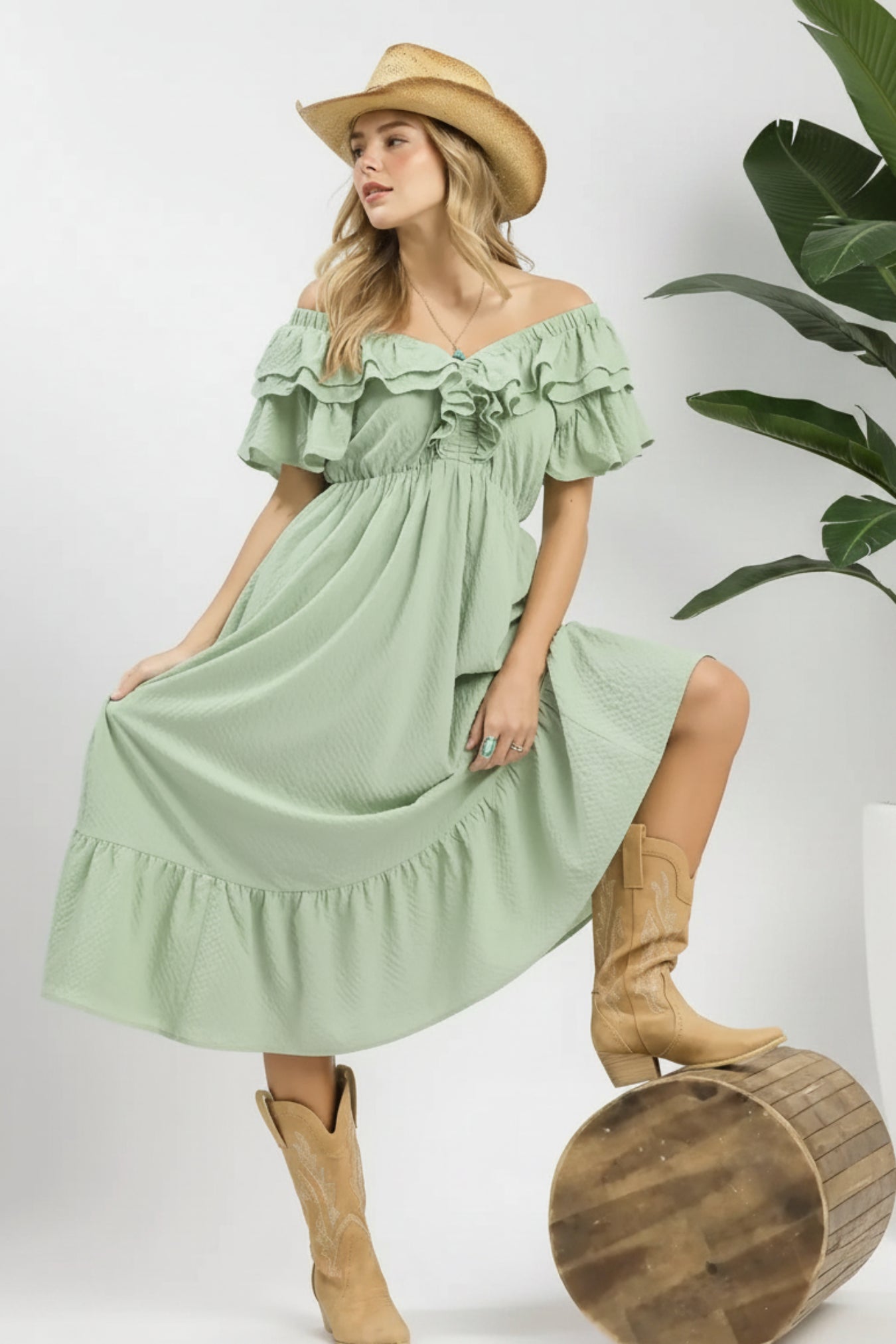 Woman wearing a light green dress with ruffles, tan cowboy boots, and a straw hat on a white background.