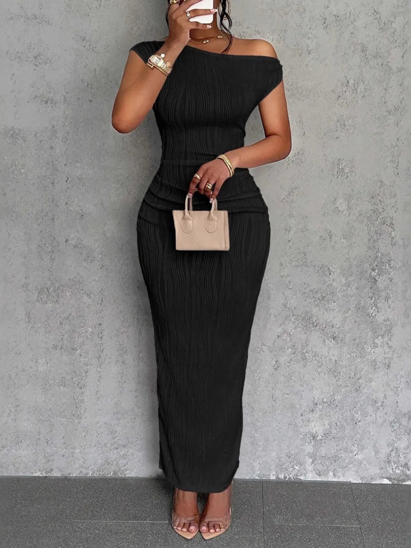 Woman wearing a black off-shoulder dress holding a beige handbag against a gray wall.