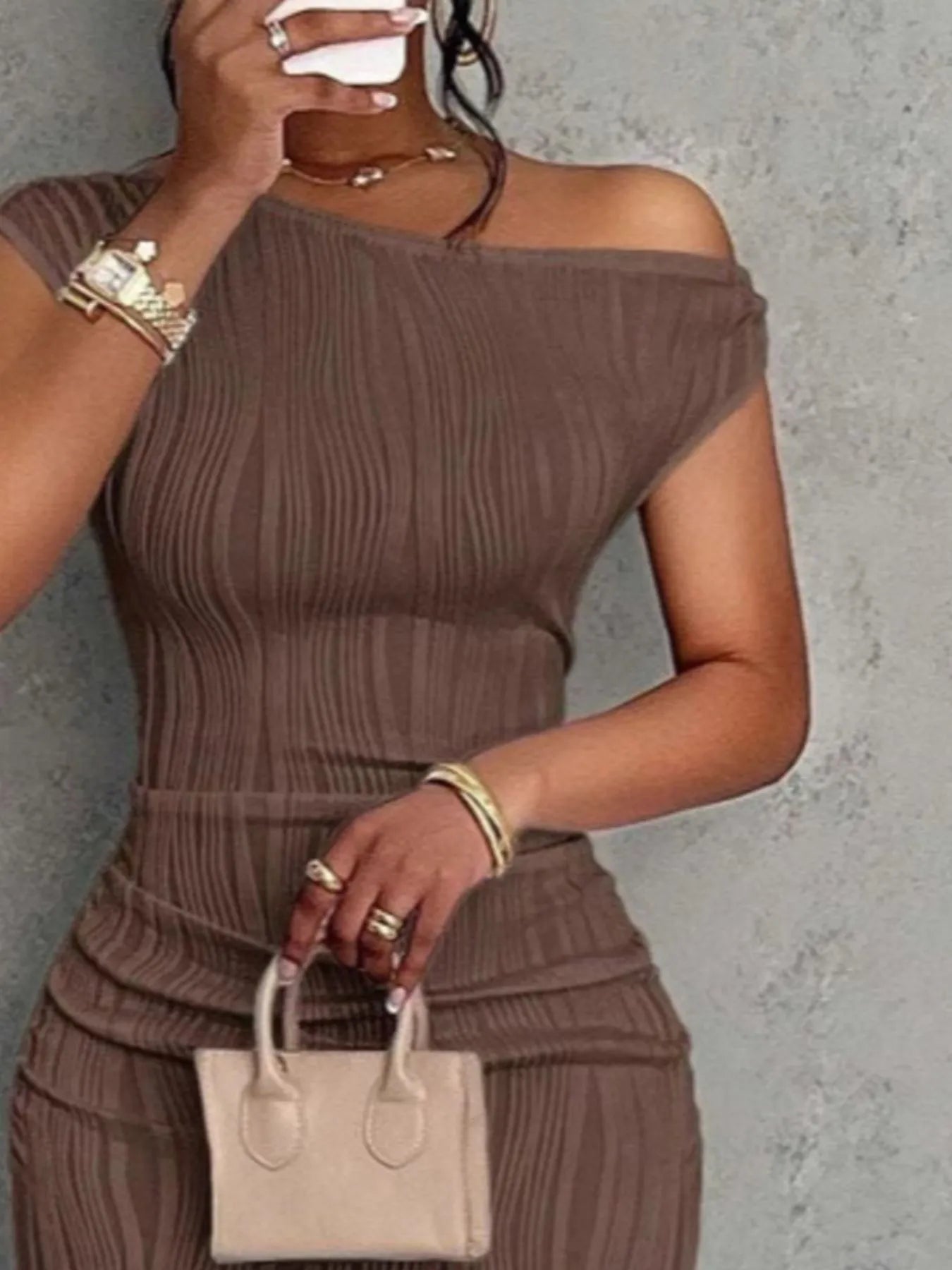 Woman in a brown ribbed outfit holding a beige handbag against a gray background