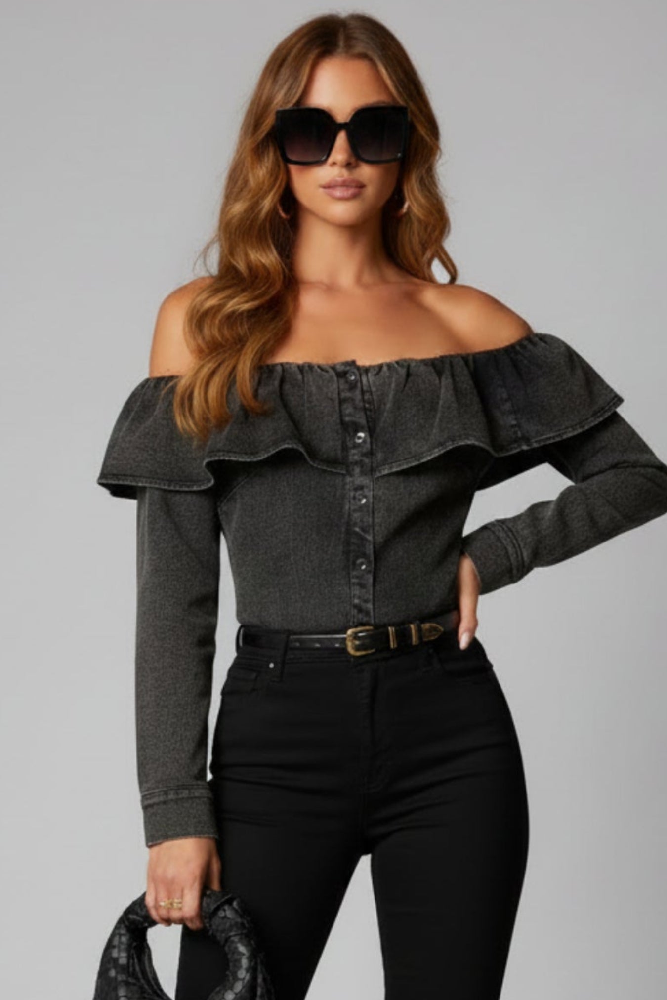 Woman wearing a black off-shoulder top and black pants with sunglasses and a handbag on a gray background