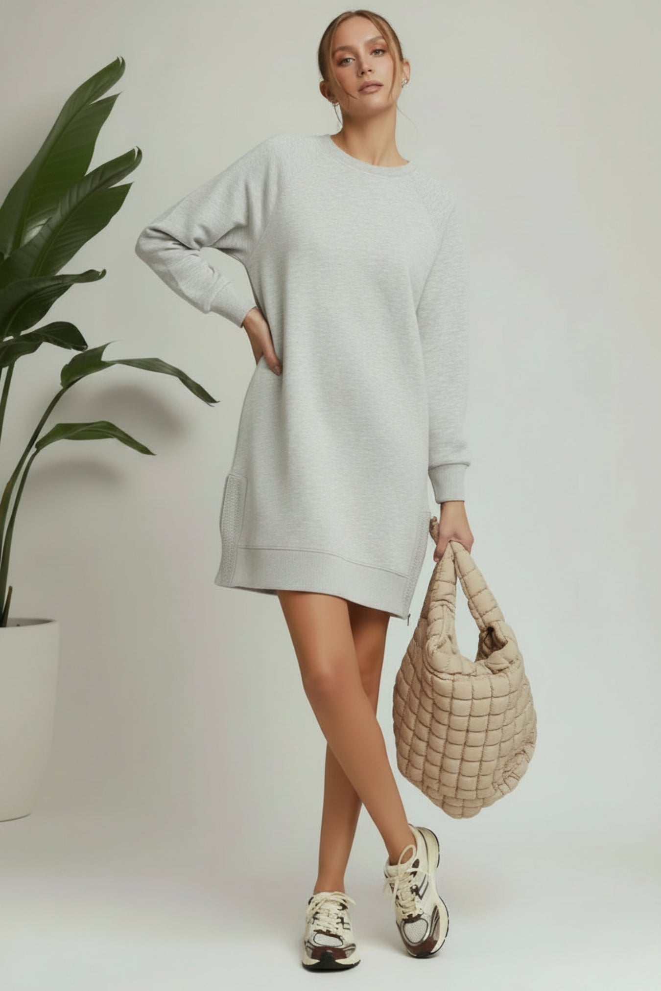Woman wearing a light gray sweater dress holding a beige woven handbag against a plain background.
