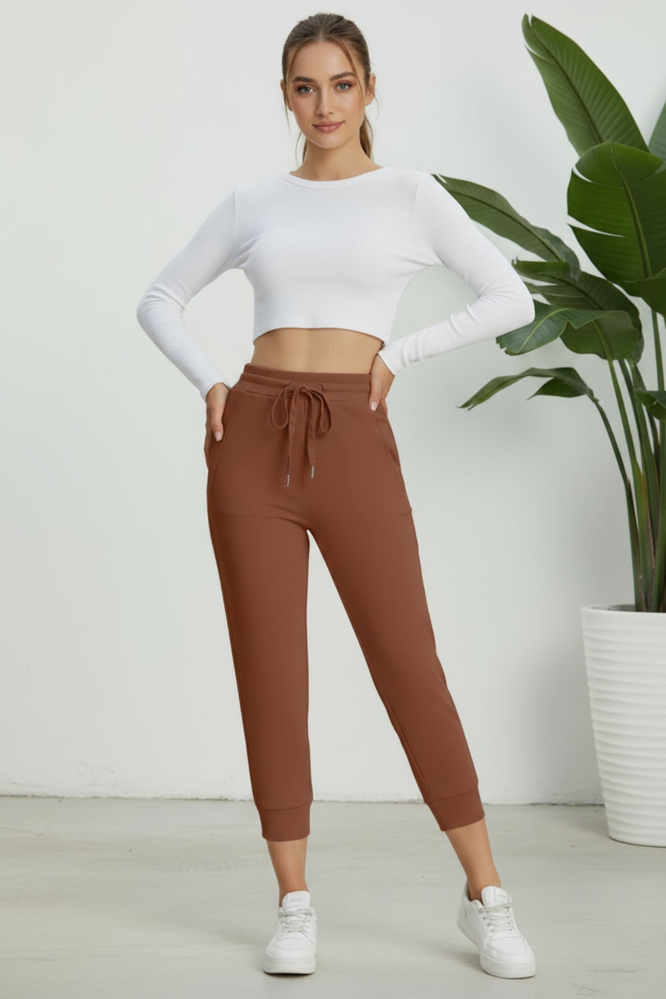 A woman modeling a white long-sleeve crop top and brown jogger pants with a drawstring waist, standing in a bright indoor setting with a potted plant in the background.