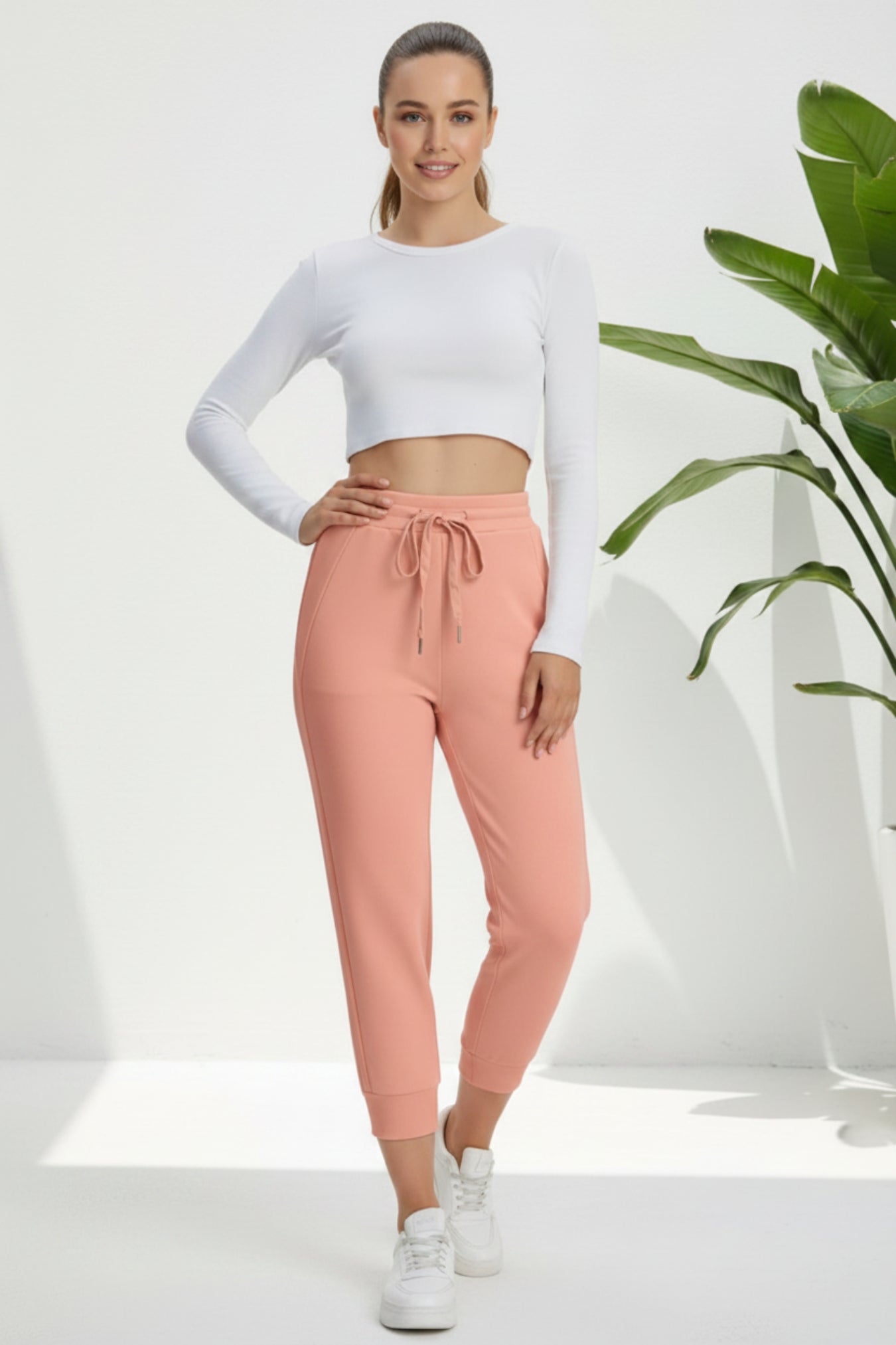 A woman modeling a white long-sleeve crop top and dusty pink jogger pants with a drawstring waist, paired with white sneakers, in a bright studio setting with a green plant in the background.