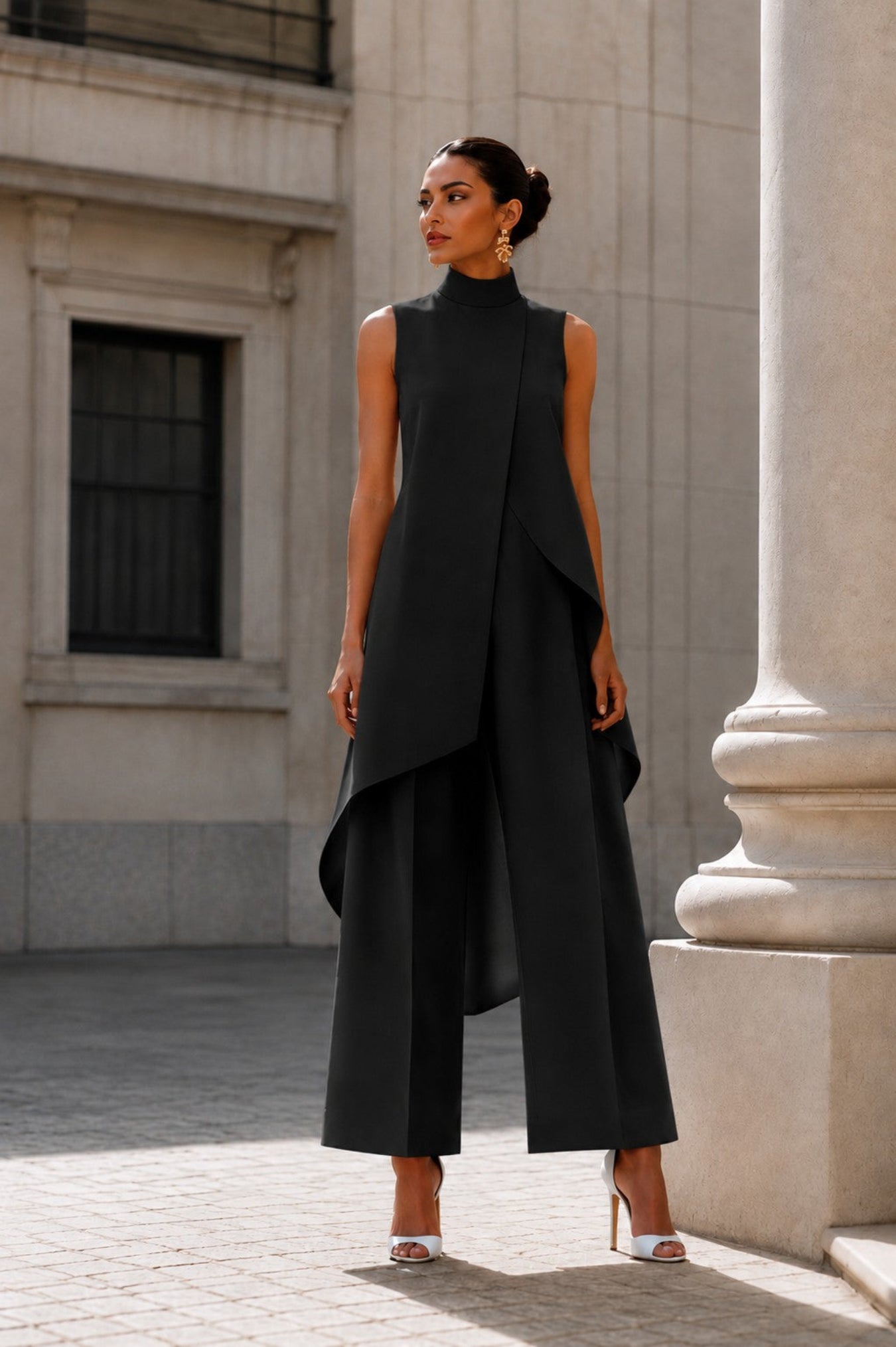 A woman stands outdoors in front of a stone building and large column, wearing a black sleeveless high-neck draped outfit with wide-leg pants and white heeled sandals.