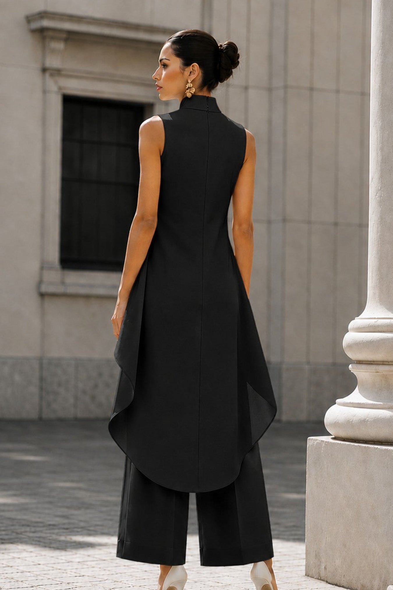 A woman wearing a black sleeveless high-low tunic top and matching wide-leg trousers, photographed from the back in an outdoor urban setting with a stone building and column in the background.