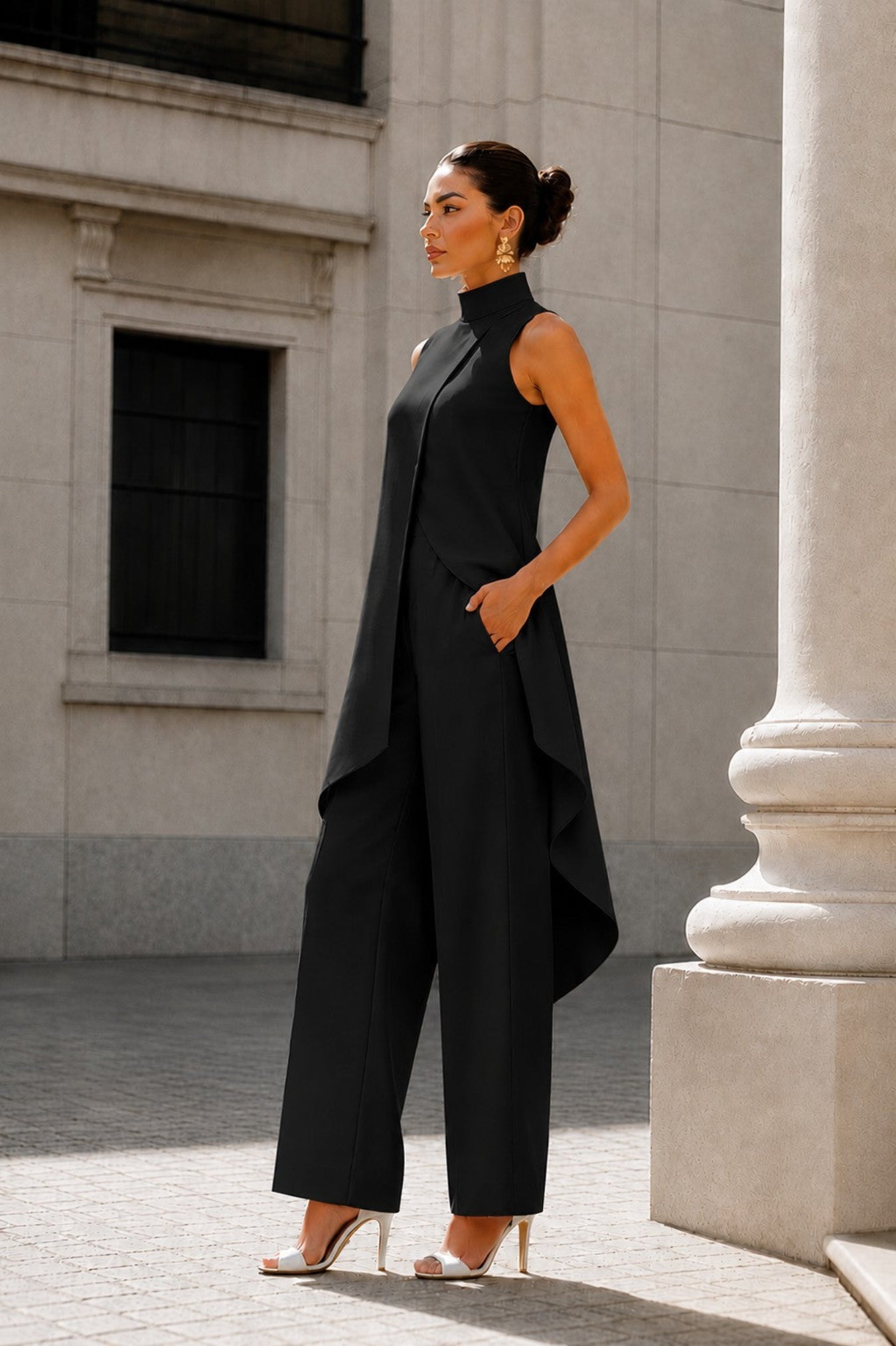 A woman models a black sleeveless high-neck jumpsuit with draped asymmetrical layers and wide legs, paired with white high-heeled sandals, standing outdoors in front of a light stone building.
