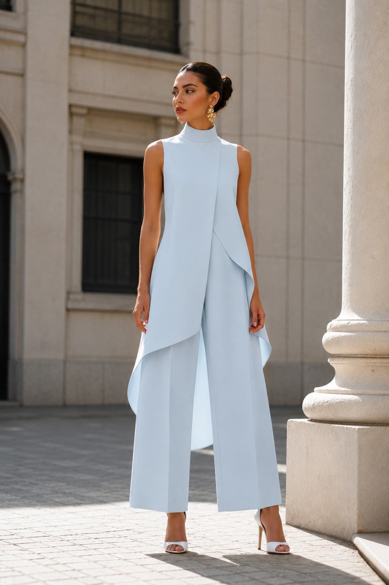 A woman models a light blue sleeveless high-neck jumpsuit with draped asymmetrical layers and wide legs, standing outdoors in front of a stone building.