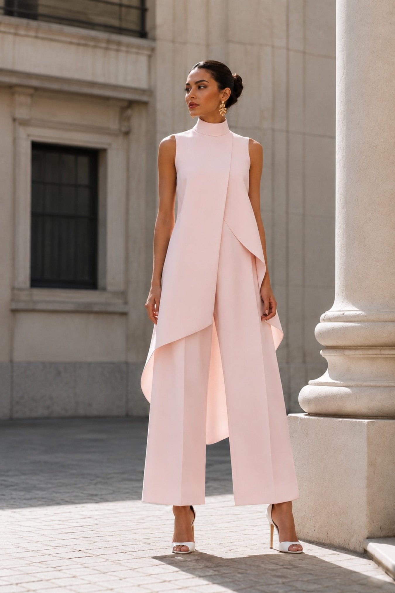 A woman models a sleeveless, high-neck, light pink jumpsuit with draped asymmetrical layers and wide legs, standing outdoors in front of a stone building with a large column.
