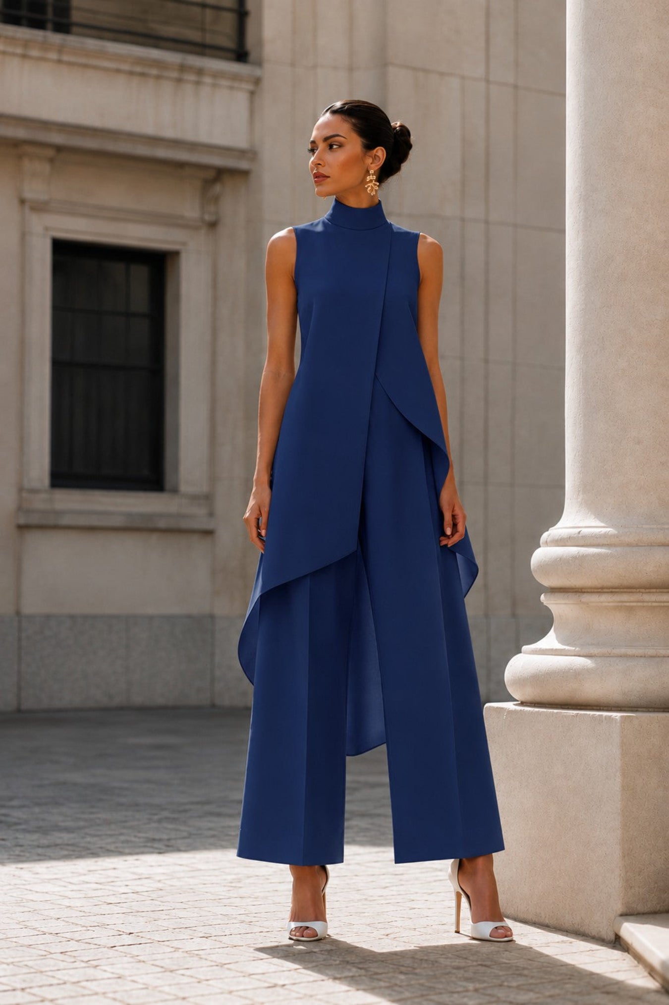 A woman models a sleeveless royal blue high-neck jumpsuit with dramatic layered asymmetrical drapes and wide-leg cropped pants, styled with white high-heeled sandals in an outdoor urban setting with a stone column.