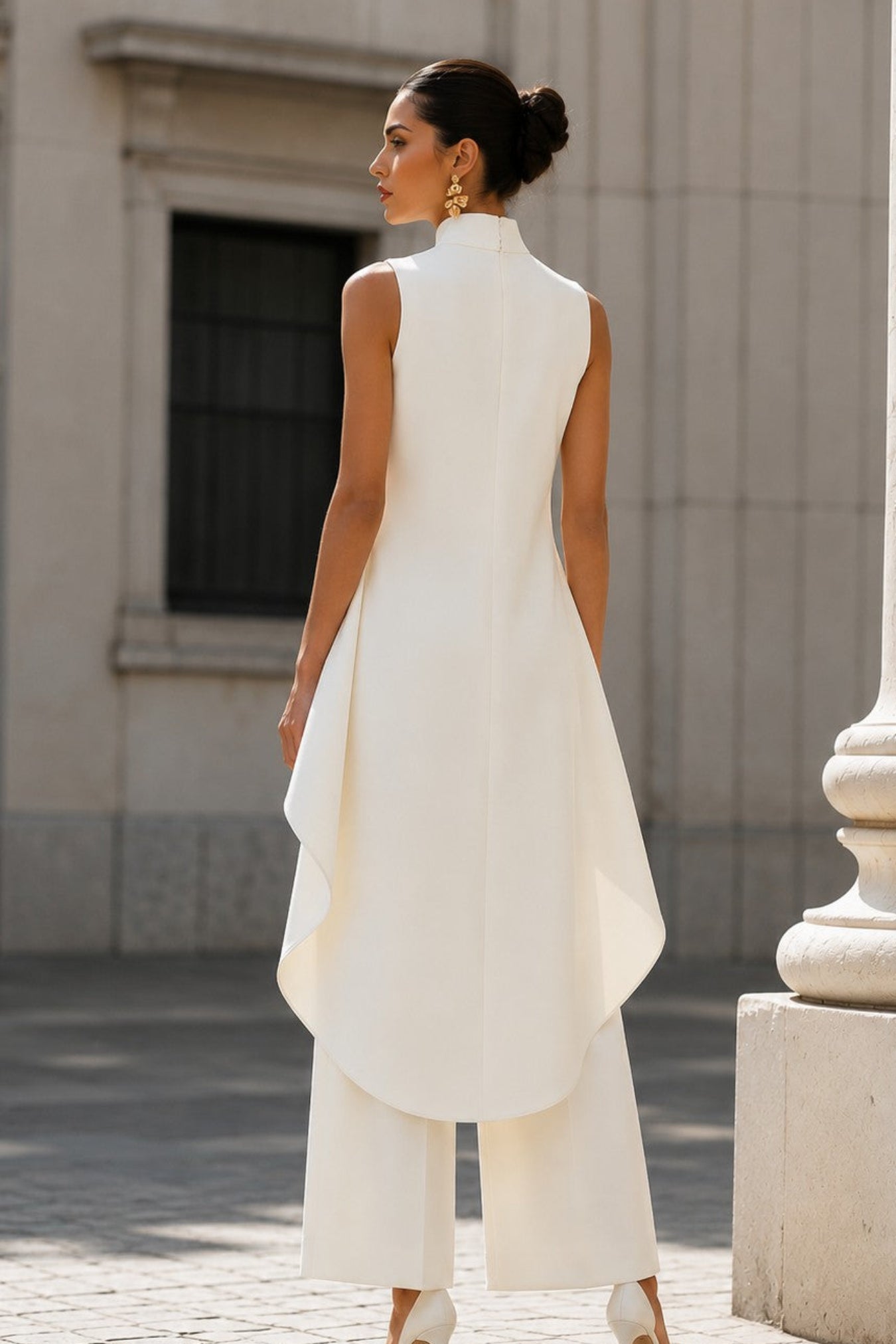 A woman wearing a white sleeveless high-neck tunic top with wide-leg cropped pants, photographed from behind in an outdoor urban setting with stone architecture.