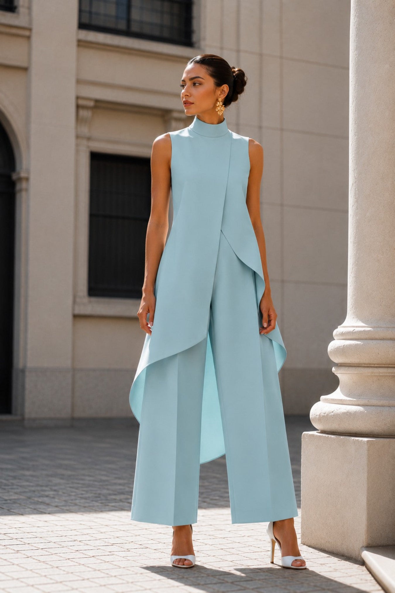 A woman models a sleeveless light blue high-neck jumpsuit with draped asymmetrical layers and wide legs, standing outdoors in front of a beige stone building.