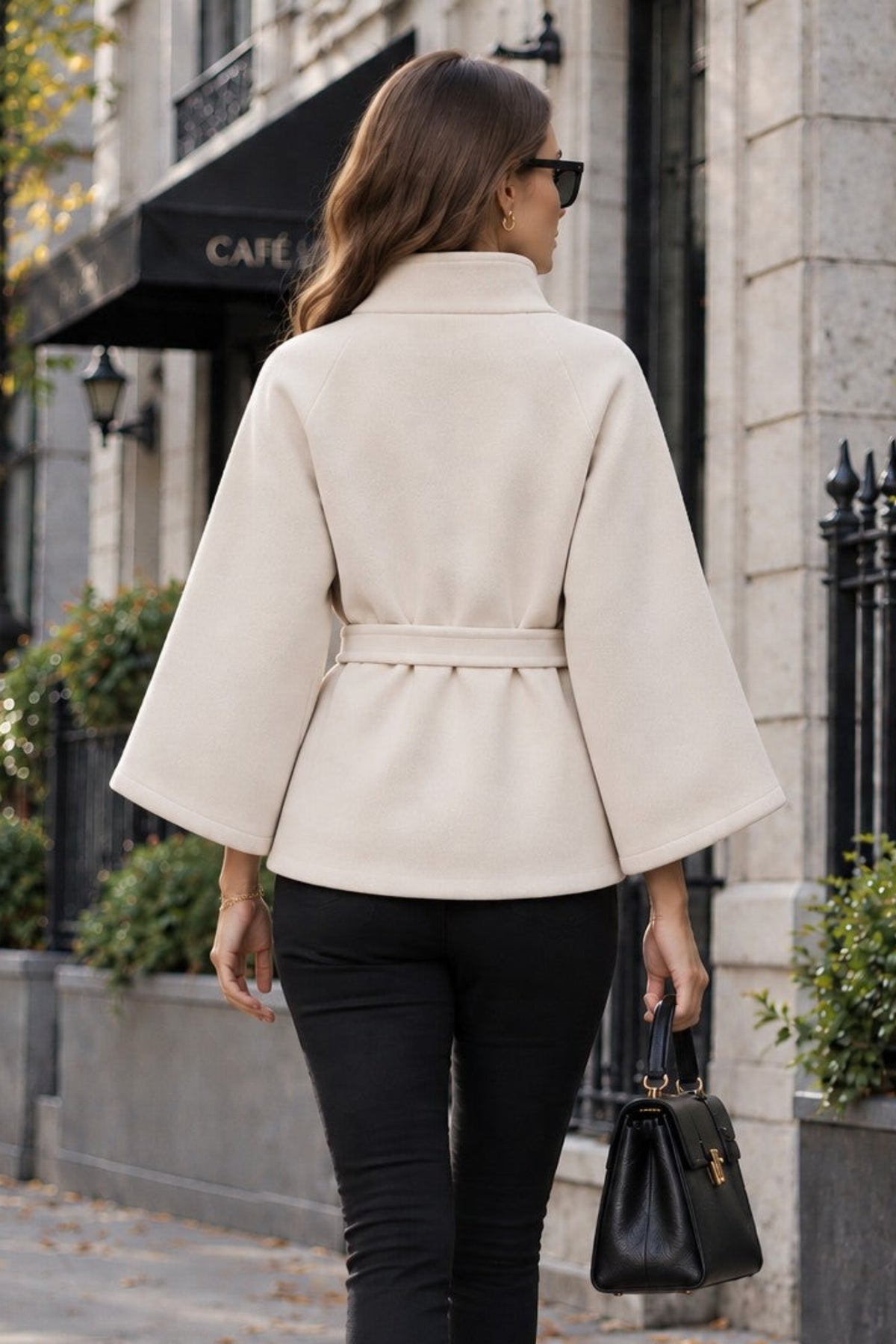 A woman viewed from behind walking on a city sidewalk, wearing a cream-colored belted cape jacket with wide sleeves, black skinny pants, black ankle boots, and carrying a black handbag.