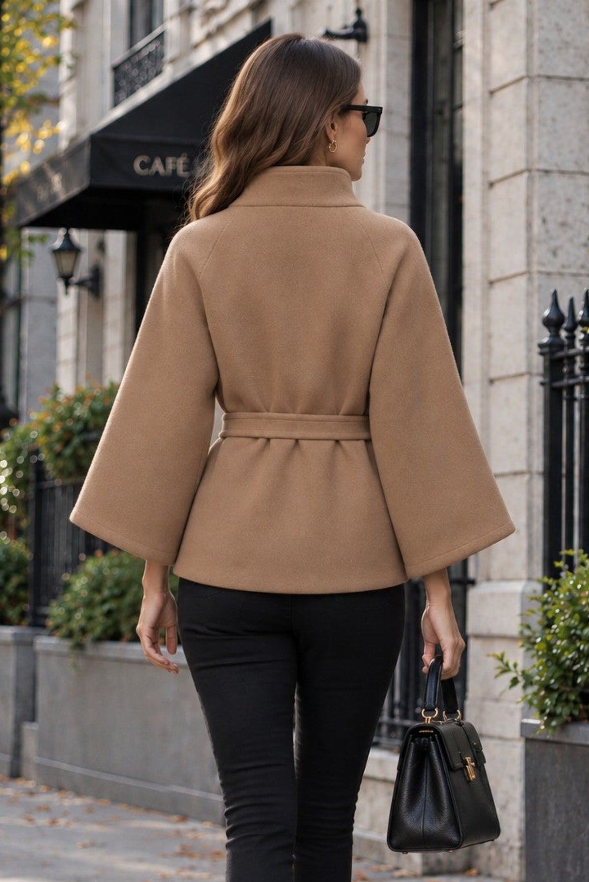 A woman wearing a camel beige belted cape jacket, black skinny pants, and black ankle boots walks down a city sidewalk, holding a black handbag.