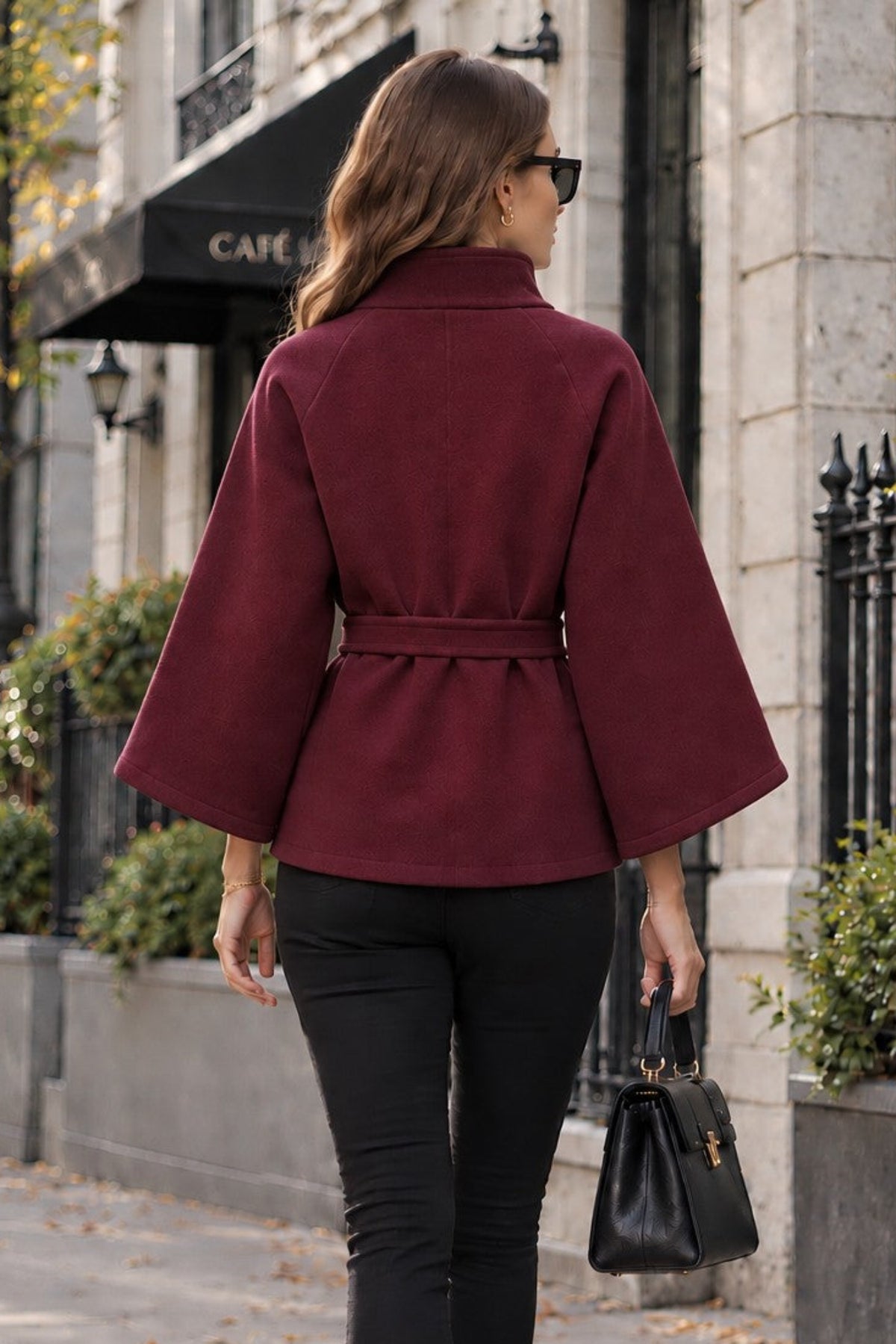 A woman viewed from behind walking on a city sidewalk, wearing a burgundy wrap-style jacket with wide sleeves, black pants, black ankle boots, and carrying a black handbag.