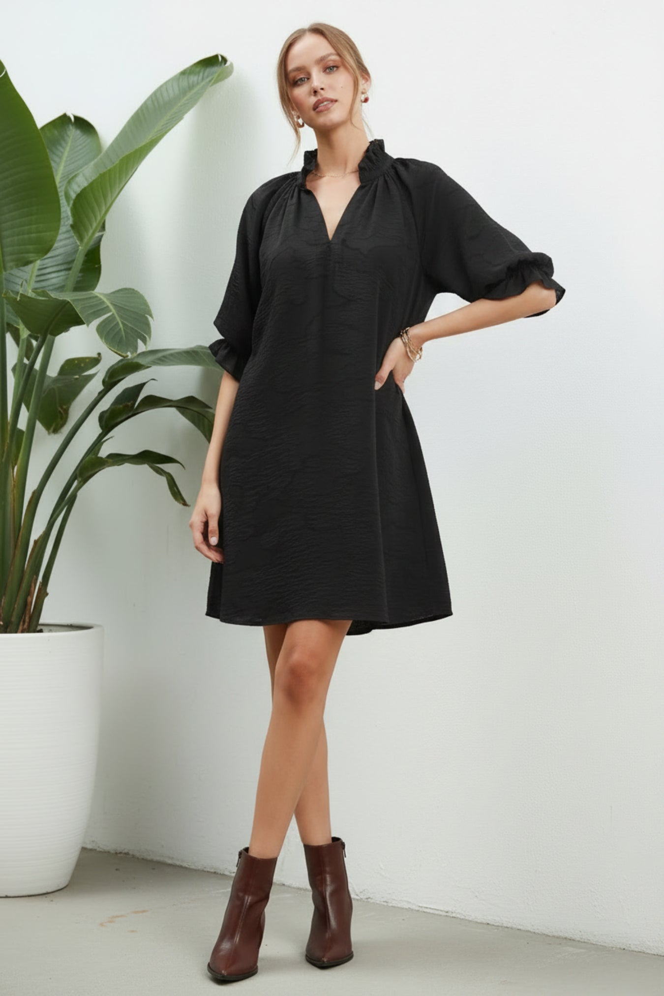 A woman modeling a loose black short-sleeve mini dress with a V-neck, paired with dark brown ankle boots, against a white background next to a plant.