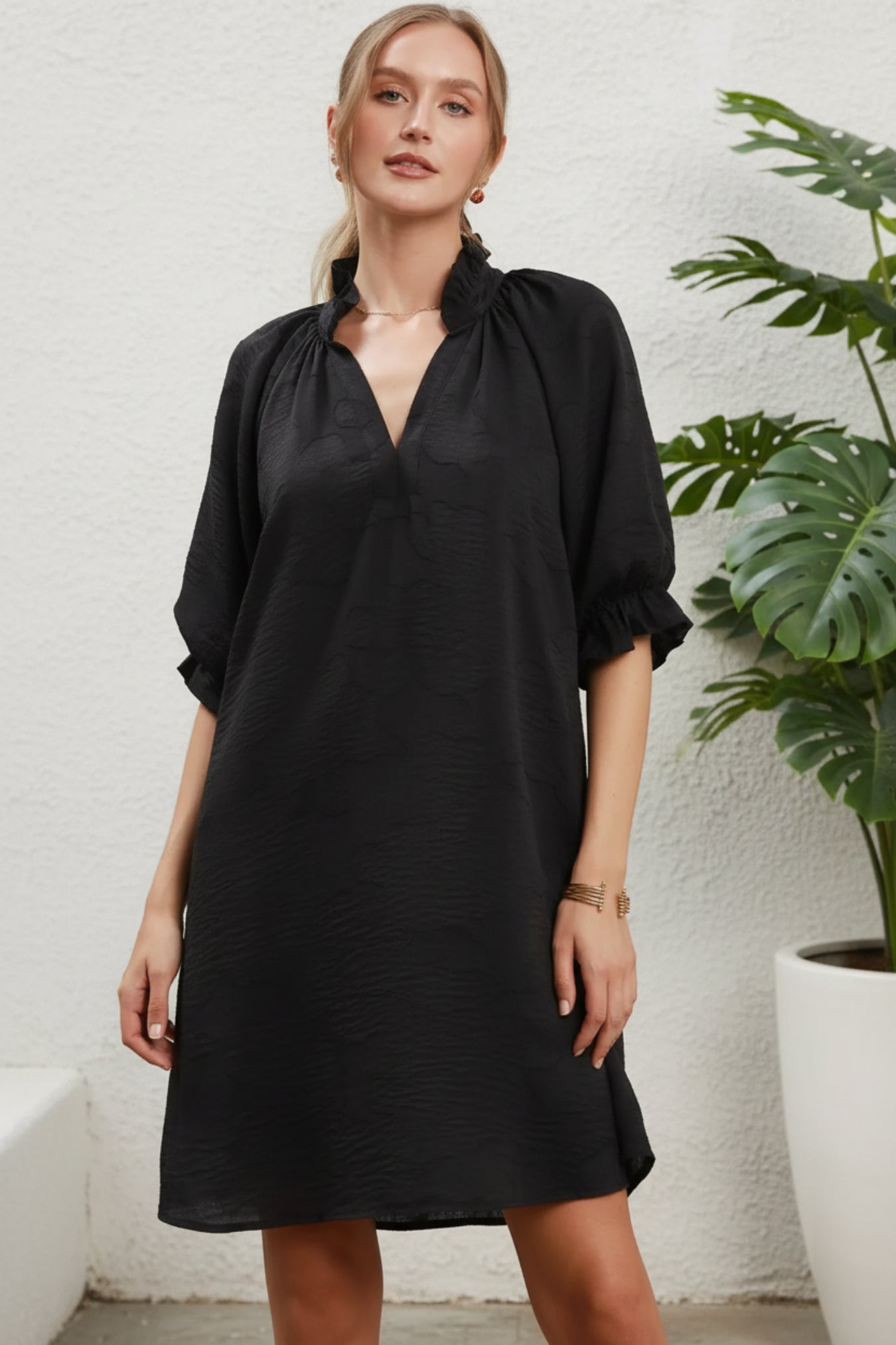A woman modeling a loose black short-sleeve tunic dress with a gathered high neckline and ruffled sleeves, posed against a white background next to a plant.