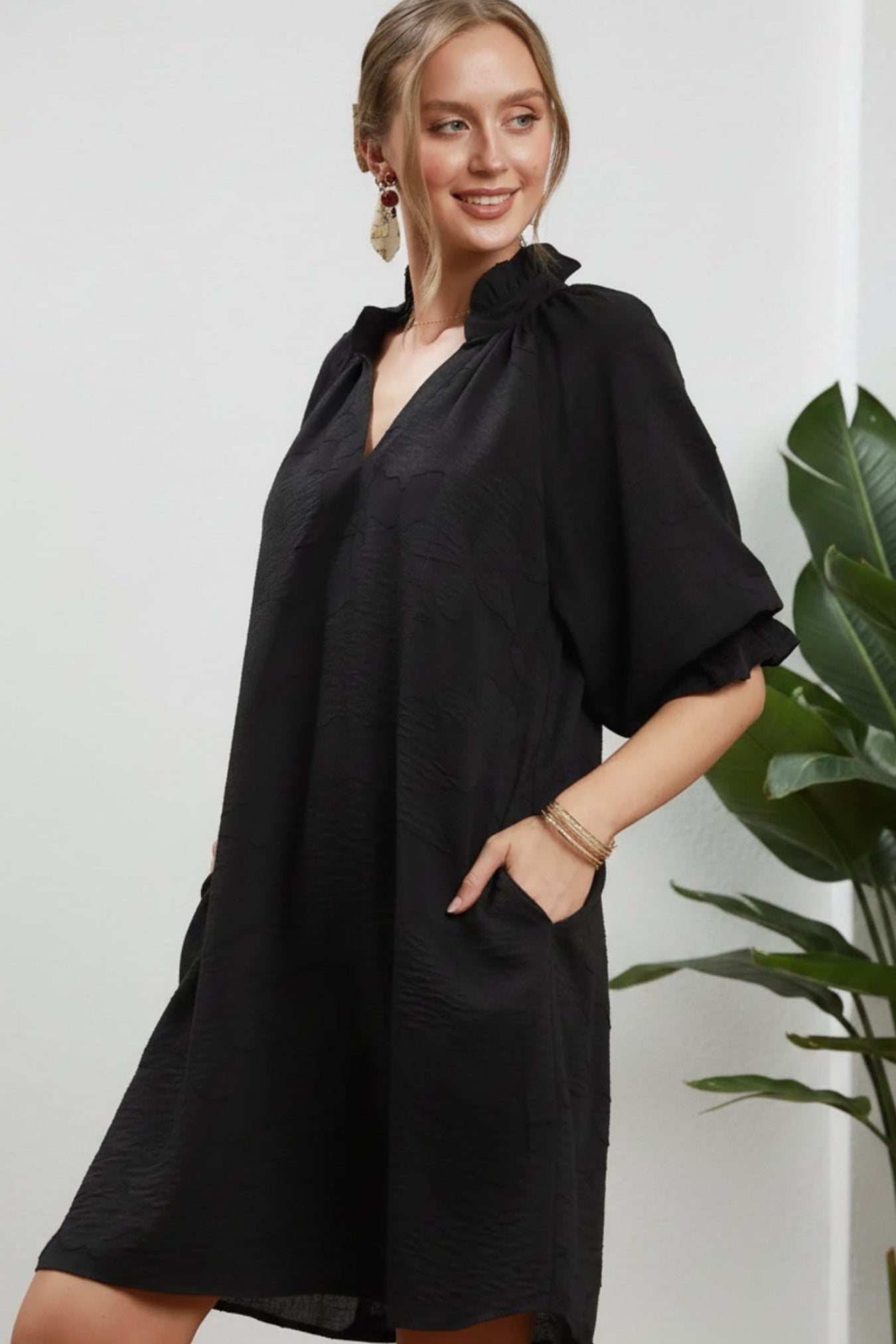 A woman modeling a loose black short-sleeve dress with a gathered high neckline and side pockets, posed against a white background.