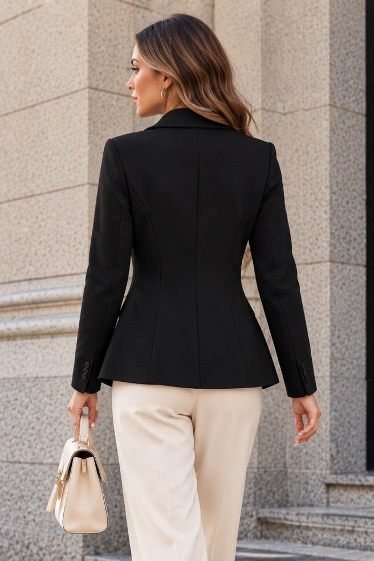 A woman photographed from behind wearing a tailored black blazer and cream wide-leg trousers, standing on stone steps outside a building and holding a small beige handbag.