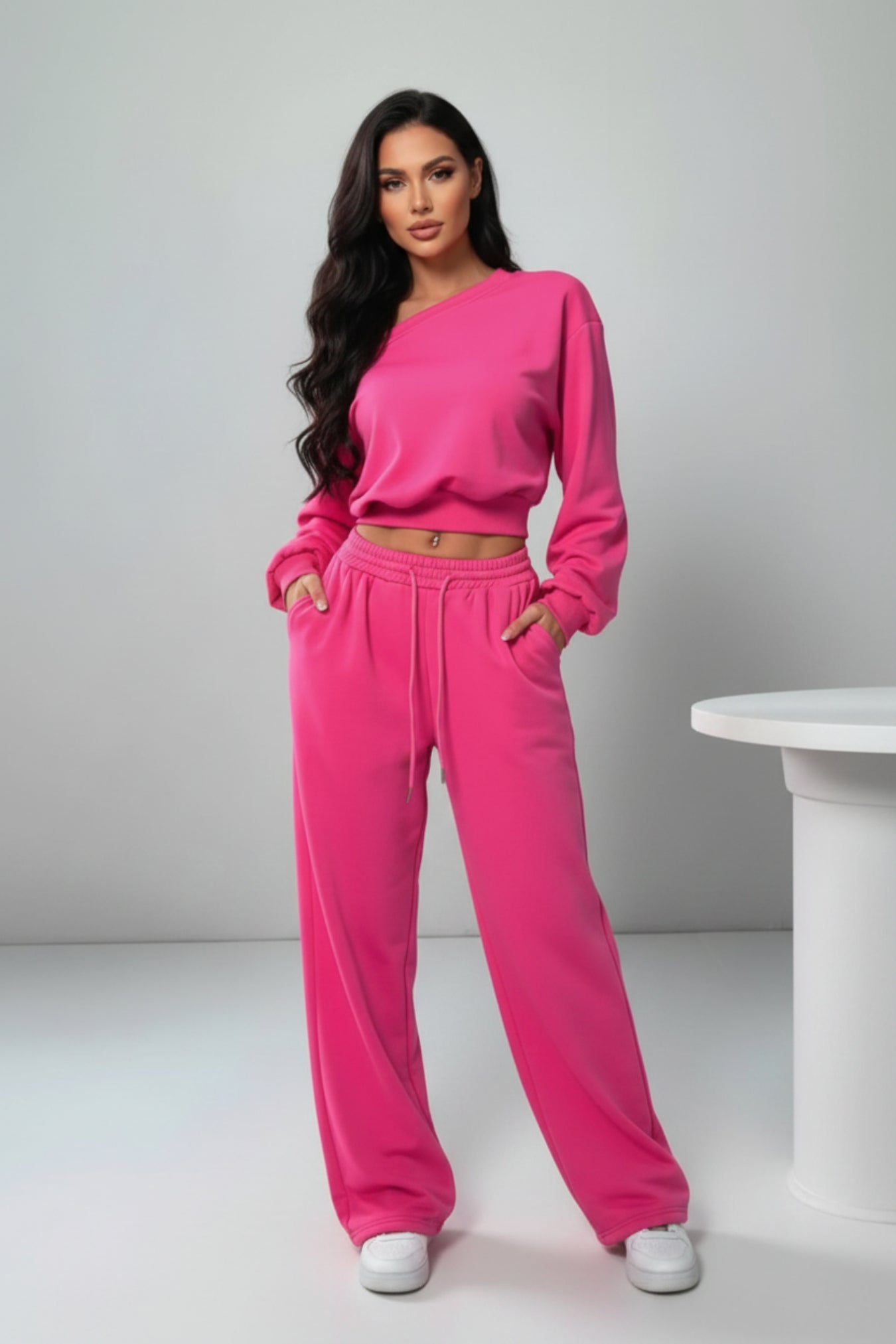 A woman modeling a bright pink two-piece lounge set consisting of a cropped long-sleeve sweatshirt and matching wide-leg sweatpants, paired with white sneakers, in a minimalist studio setting.