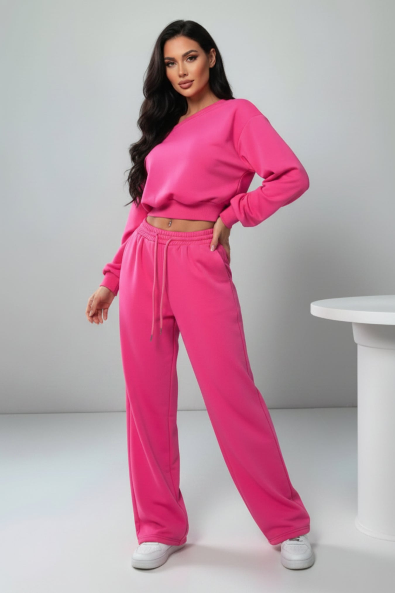 A woman modeling a bright pink two-piece lounge set consisting of a long-sleeve crop top and matching wide-leg sweatpants, paired with white sneakers, in a minimalist studio setting.