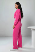 A woman modeling a bright pink two-piece lounge set consisting of a cropped long-sleeve sweatshirt and matching wide-leg sweatpants, paired with white sneakers, in a studio setting.