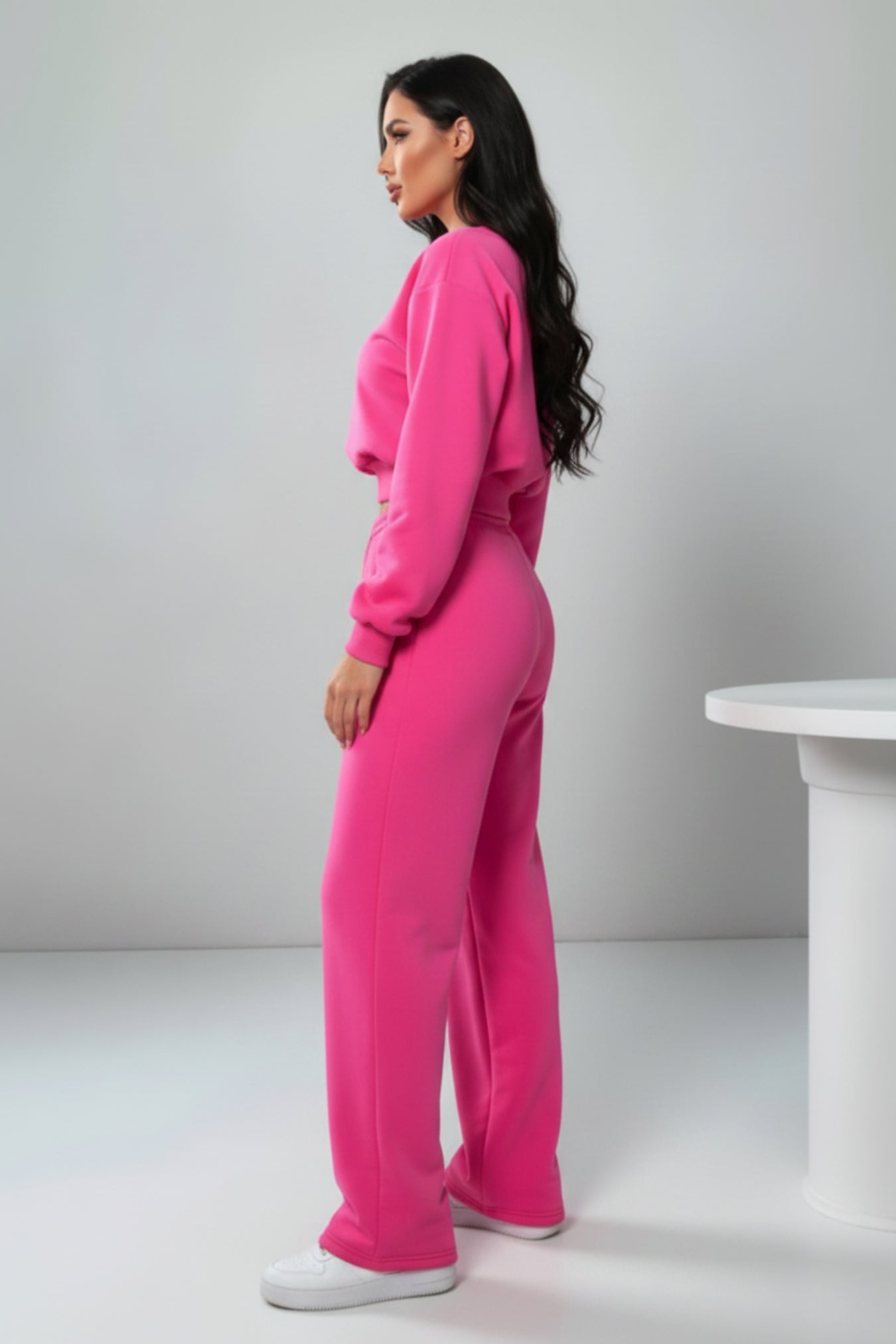 A woman modeling a bright pink two-piece lounge set consisting of a cropped long-sleeve sweatshirt and matching wide-leg sweatpants, paired with white sneakers, in a studio setting.
