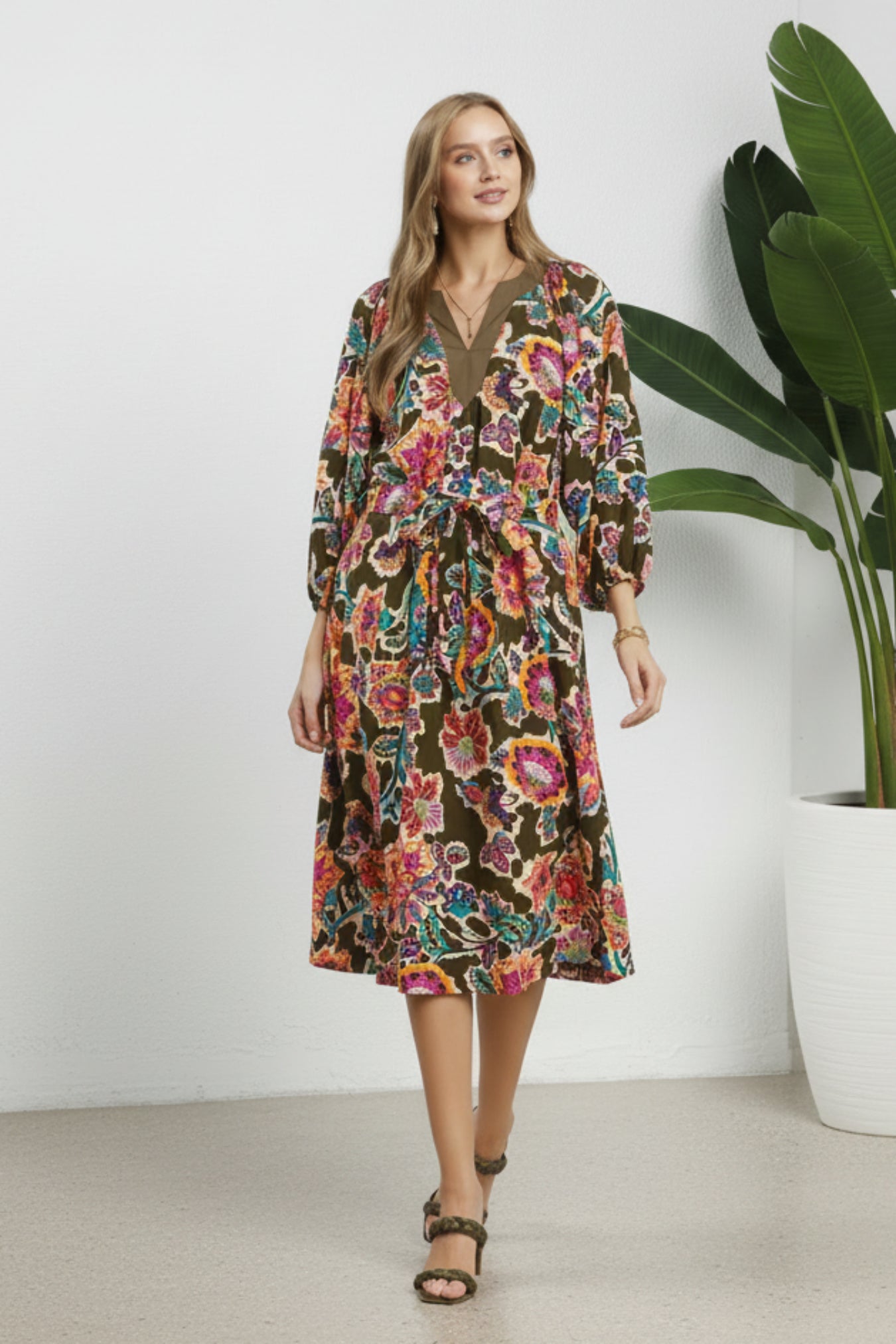 Woman wearing a colorful floral dress on a white background next to a plant
